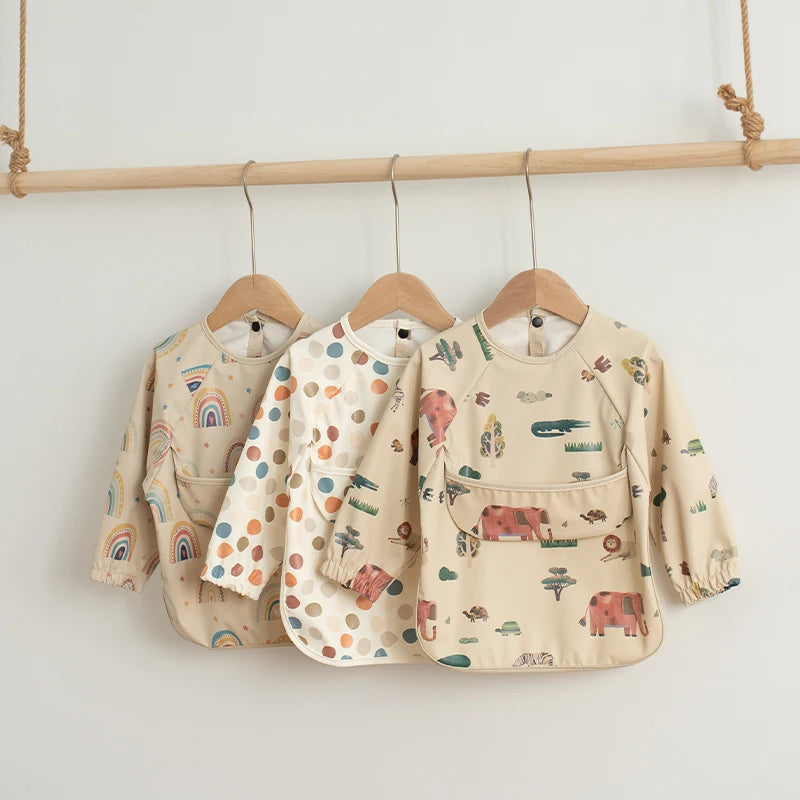 A beige waterproof long-sleeve art smock for toddlers with various nature and animal prints hanging on a wooden rack.