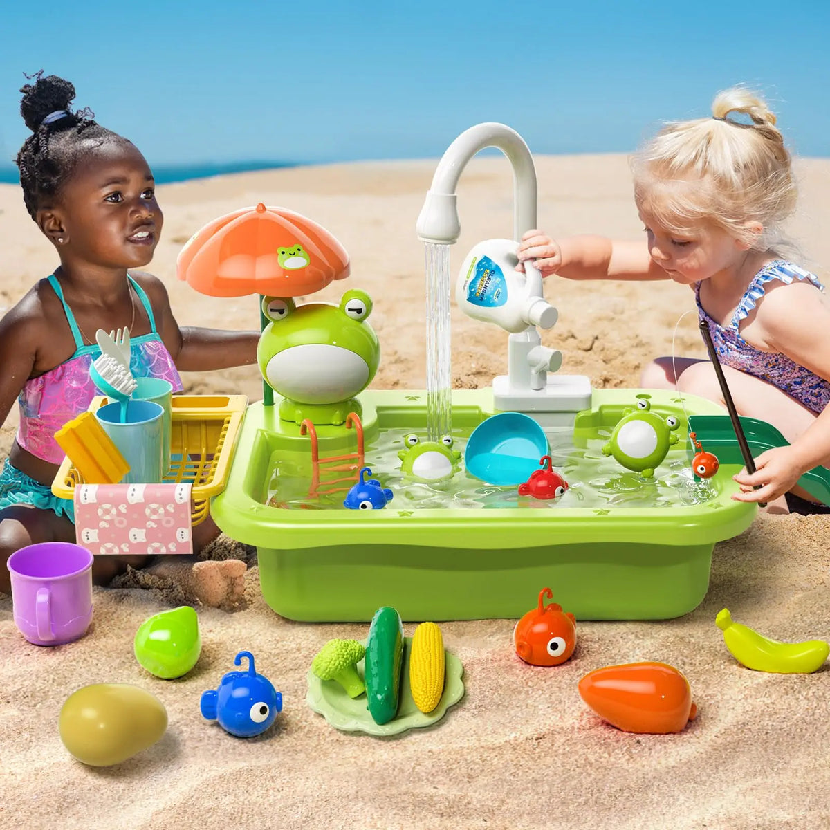 Two toddlers play with a green electric kitchen sink set featuring running water and various fruit-shaped toys on a sandy bea