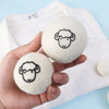 Two white wool dryer balls with black line drawings of sheep faces are held in a hand against a white cloth background.