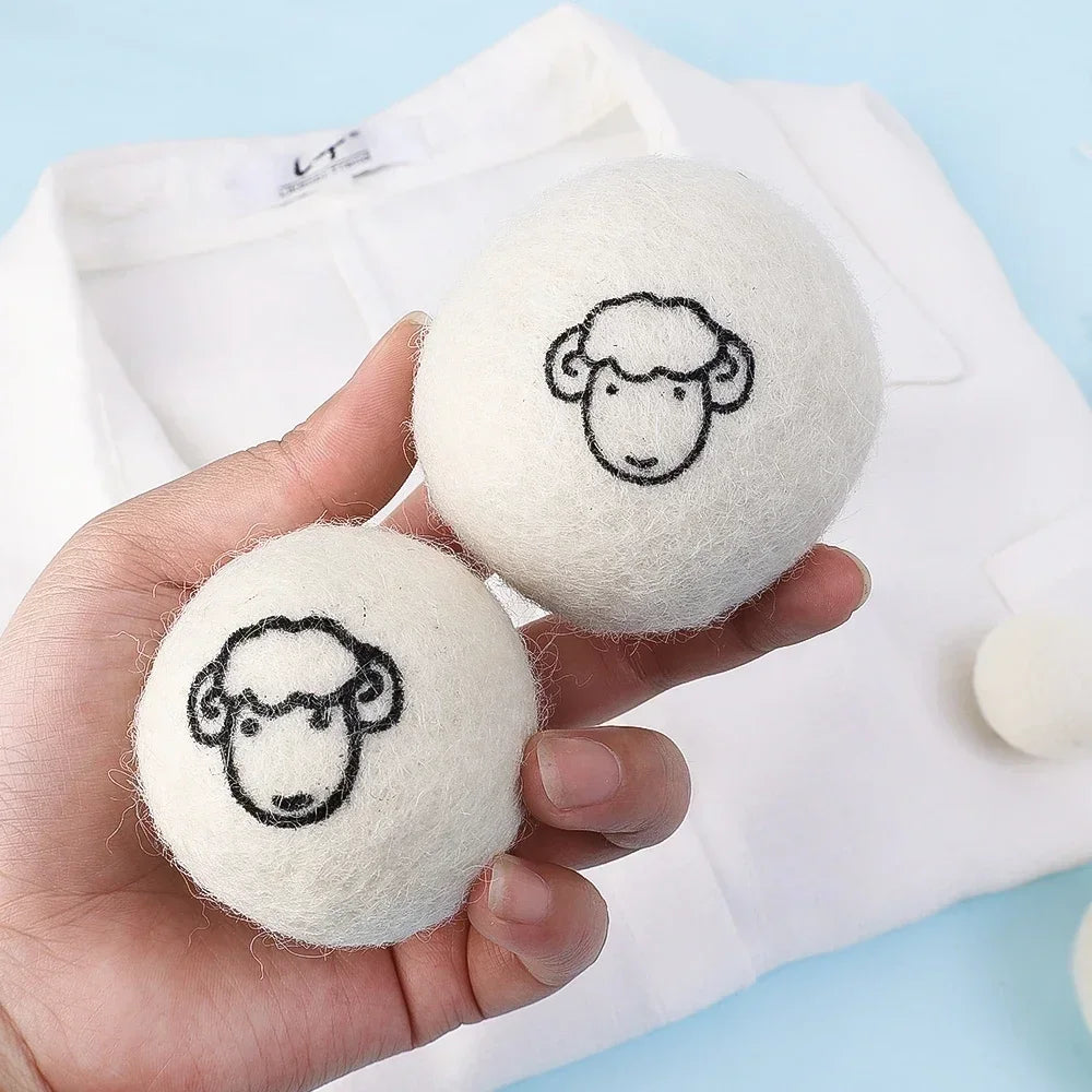 Two white wool dryer balls with black line drawings of sheep faces are held in a hand against a white cloth background.