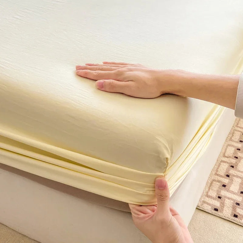 A hand is holding up a yellow fitted sheet to show its deep pocket and elastic band.