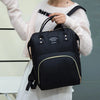 A woman wearing a cream sweater holds a black insulated canvas mummy diaper bag with a hanging hook.