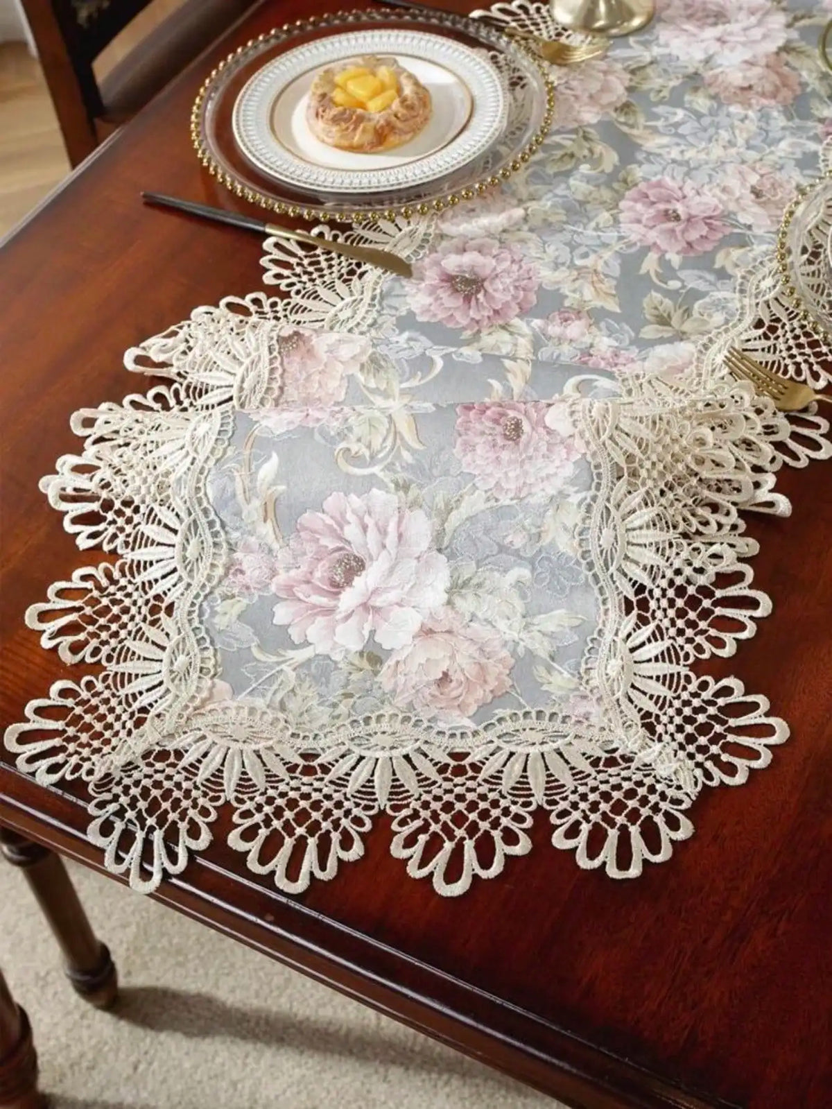 A lace table runner featuring a floral pattern in pink and gray on a dark wood table.