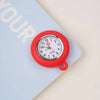 A red silicone pocket watch with a white face and black hands is pinned to a light blue notebook.