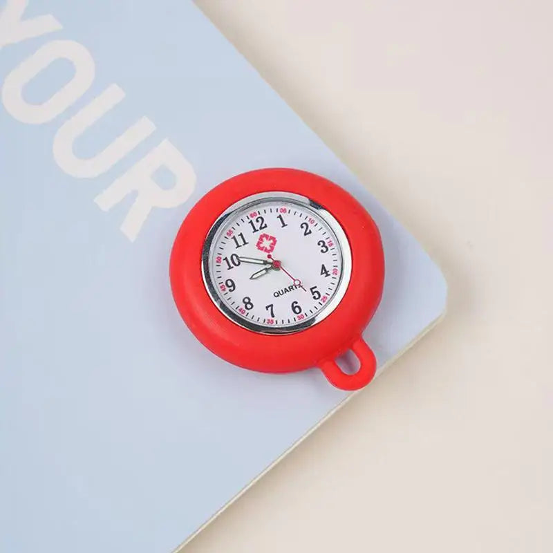 A red silicone pocket watch with a white face and black hands is pinned to a light blue notebook.