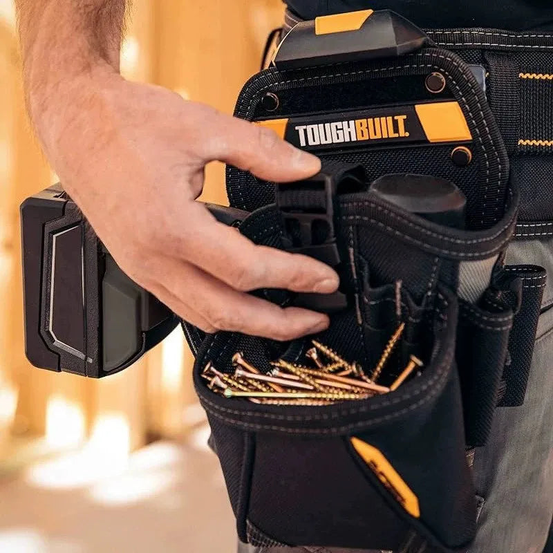 A hand is holding a black ToughBuilt tool belt with multiple pockets and gold tools.