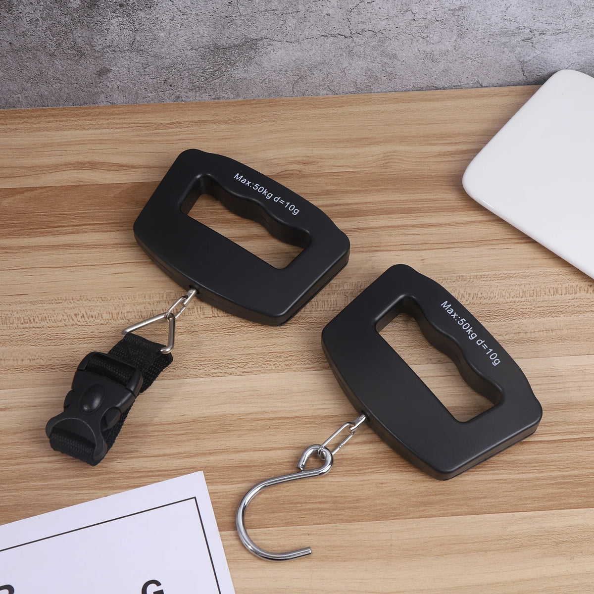 A black portable digital luggage scale is shown on a wooden surface with a hanging hook and a scale display reading Max: 50kg