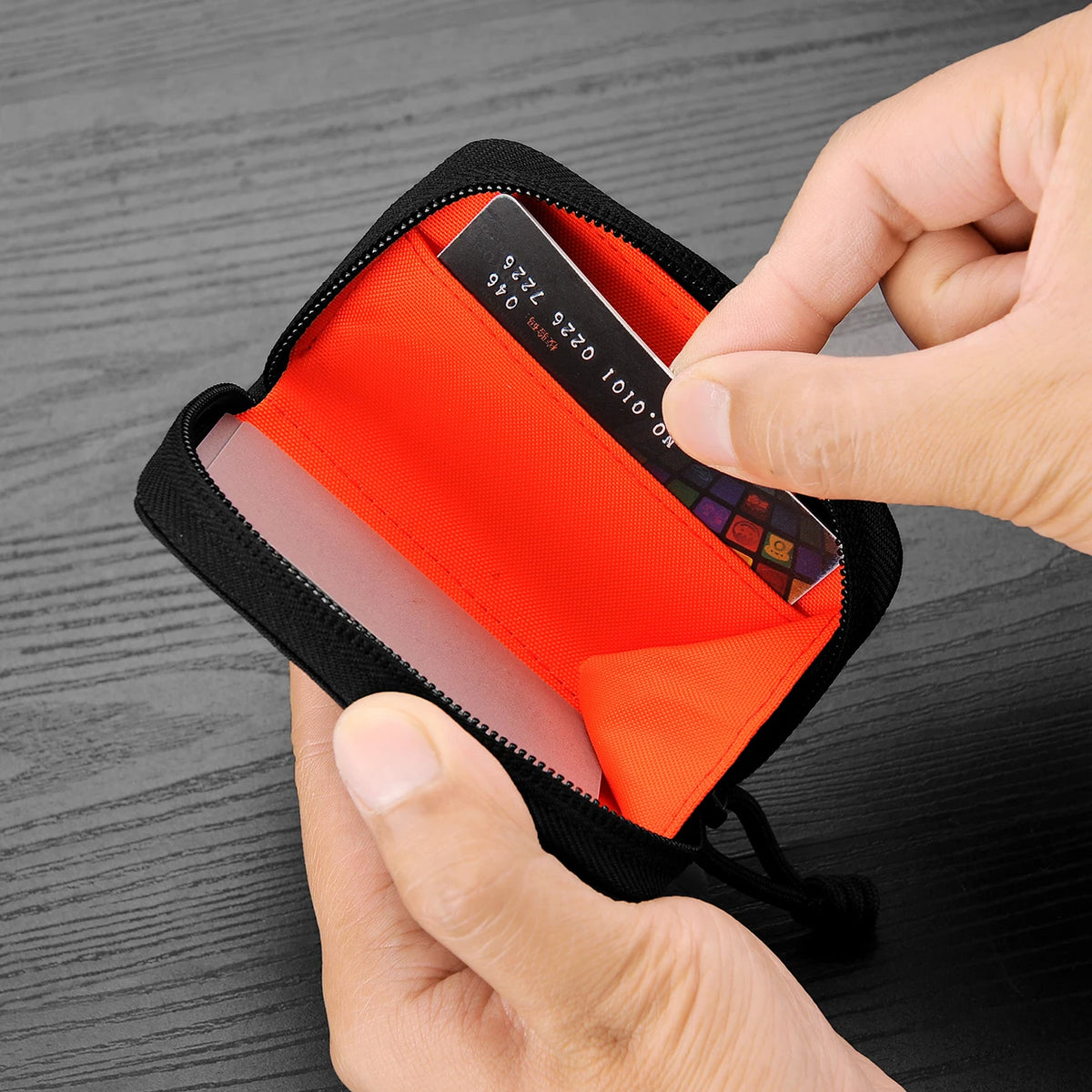 A hand is opening a black compact RFID wallet with an orange lining to reveal a credit card.