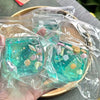 Clear plastic cube filled with blue-green glitter and various small seashells used as a stress relief fidget toy