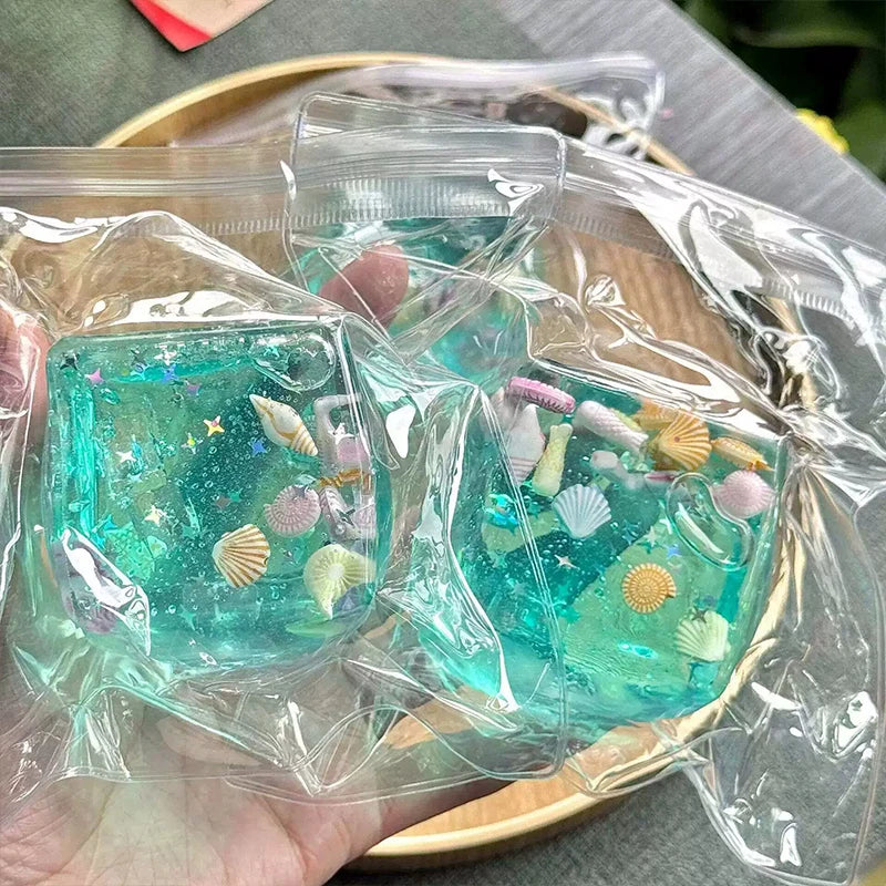 Clear plastic cube filled with blue-green glitter and various small seashells used as a stress relief fidget toy