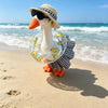 A goose costume wearing sunglasses and a straw hat standing on a sandy beach near the ocean.