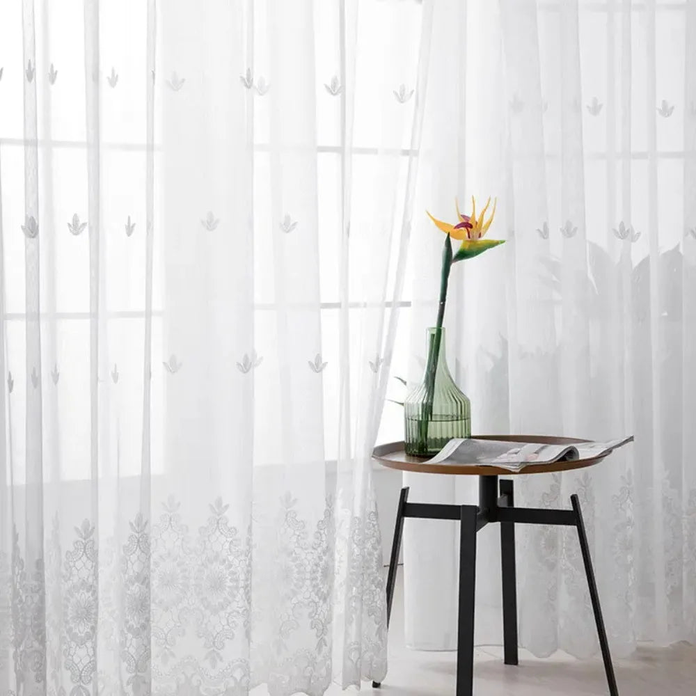 A sheer white voile curtain panel with delicate embroidery hangs in a bright window.