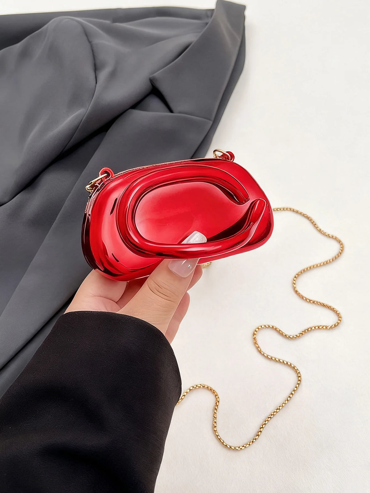 A hand holds a red mini clutch bag with gold chain strap against a gray blazer.