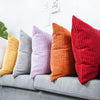 A row of nine textured corduroy cushion covers in shades of yellow, gray, lavender, orange, and red resting on a gray sofa.