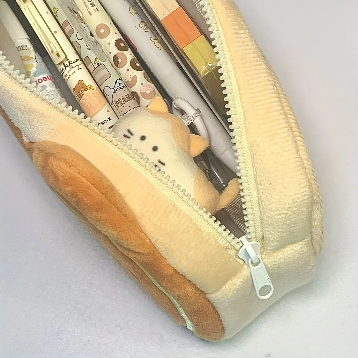 A beige and brown caramel cat plush pencil case with a zipper showing various pens and pencils inside.