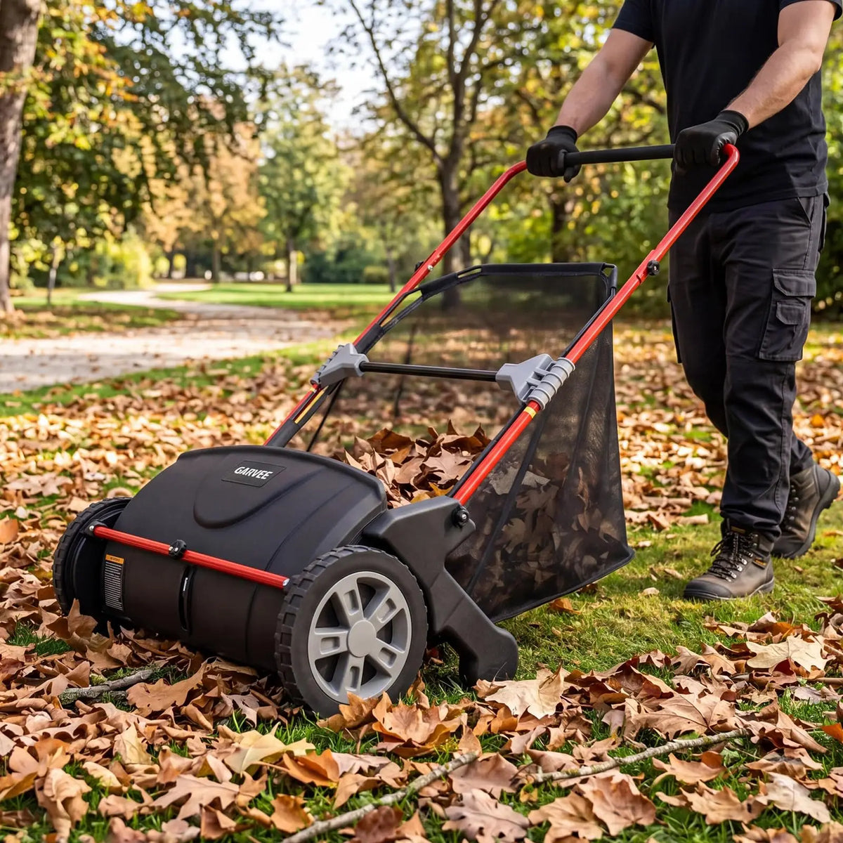 A person is operating a black push lawn sweeper with a red handle and large hopper in an outdoor park setting covered in fall