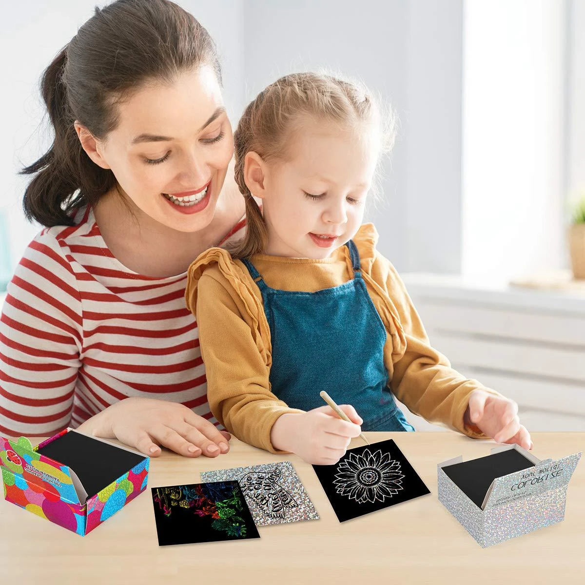 A woman and a girl are using a rainbow scratch art paper set with wooden stylus on a wooden table.