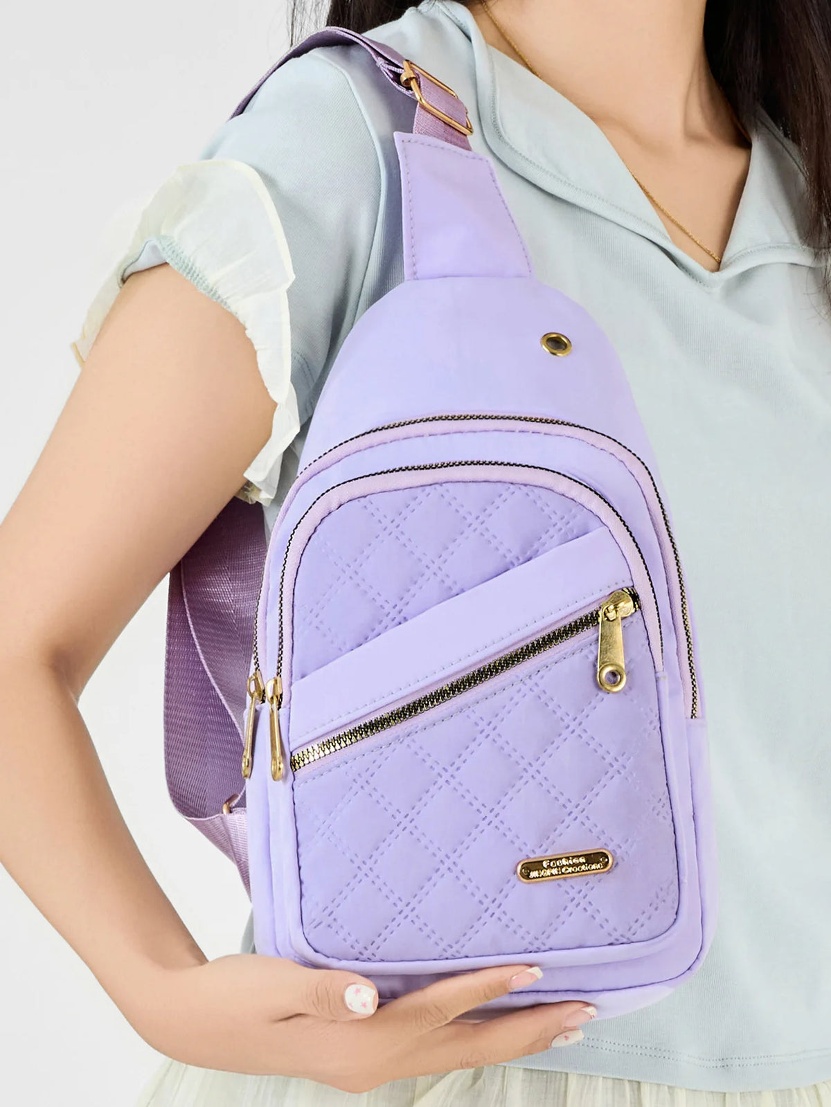 A purple quilted crossbody sling bag with a gold zipper is worn by a person in a light blue shirt.