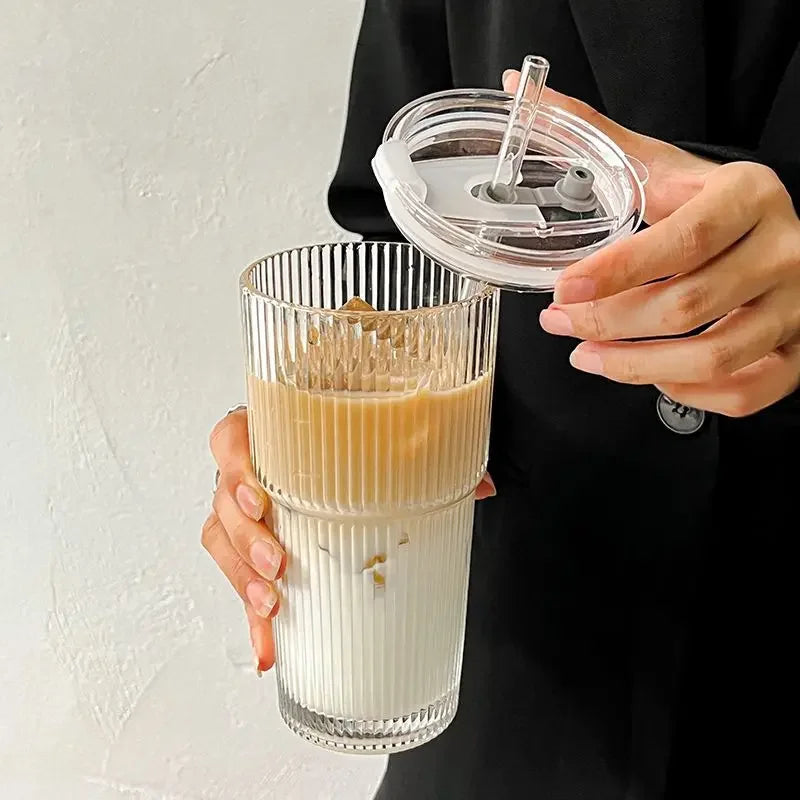 Une main tient un verre à glace rempli de boisson glacée et un couvercle transparent avec une paille sur un fond blanc.