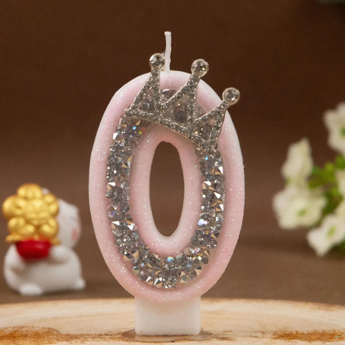 A pink number candle shaped like a zero with a rhinestone crown on top, set against a wooden surface.