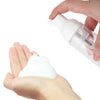 A hand is squeezing white foaming soap from a clear refillable dispenser bottle onto the palm of another hand.
