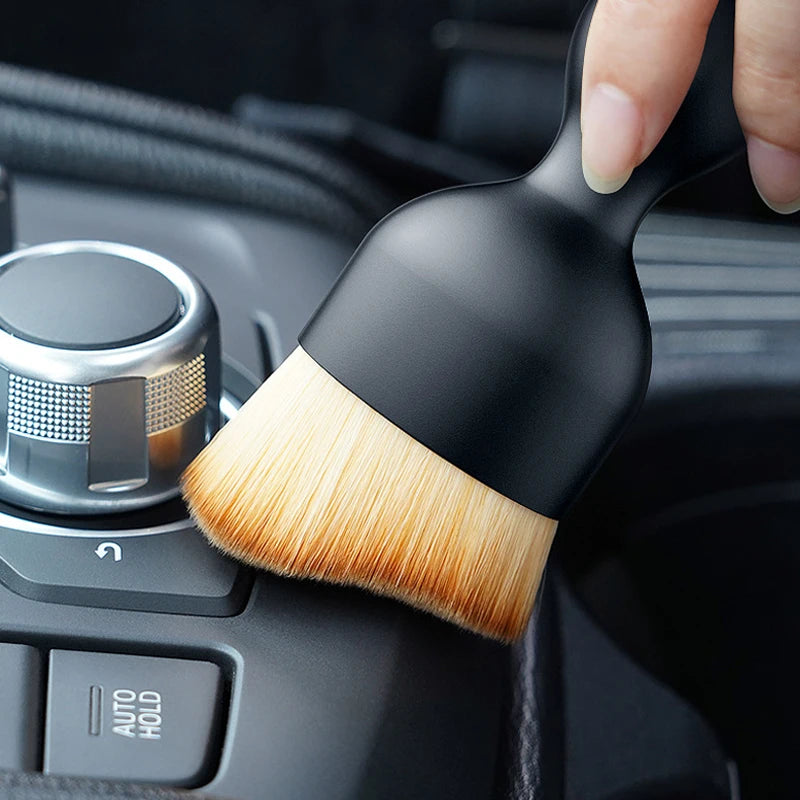 A hand holds a black cleaning brush with soft bristles, positioned near a car vent or crevice.