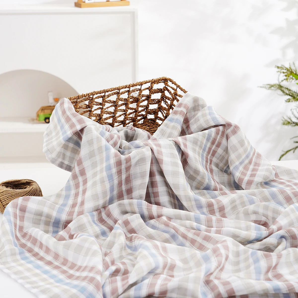 A soft, plaid muslin baby swaddle blanket draped over a woven basket in a bright, airy setting.
