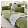 A solid olive green chenille sofa cover with light gray pillows and a matching throw blanket on a light carpet.