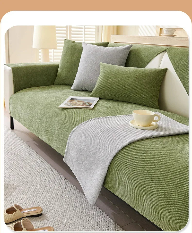 A solid olive green chenille sofa cover with light gray pillows and a matching throw blanket on a light carpet.