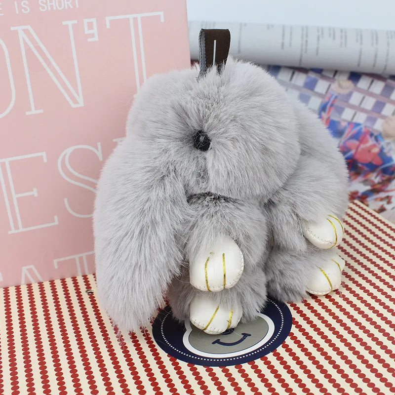 A fluffy gray bunny keychain with a pom pom tail resting on a patterned surface.