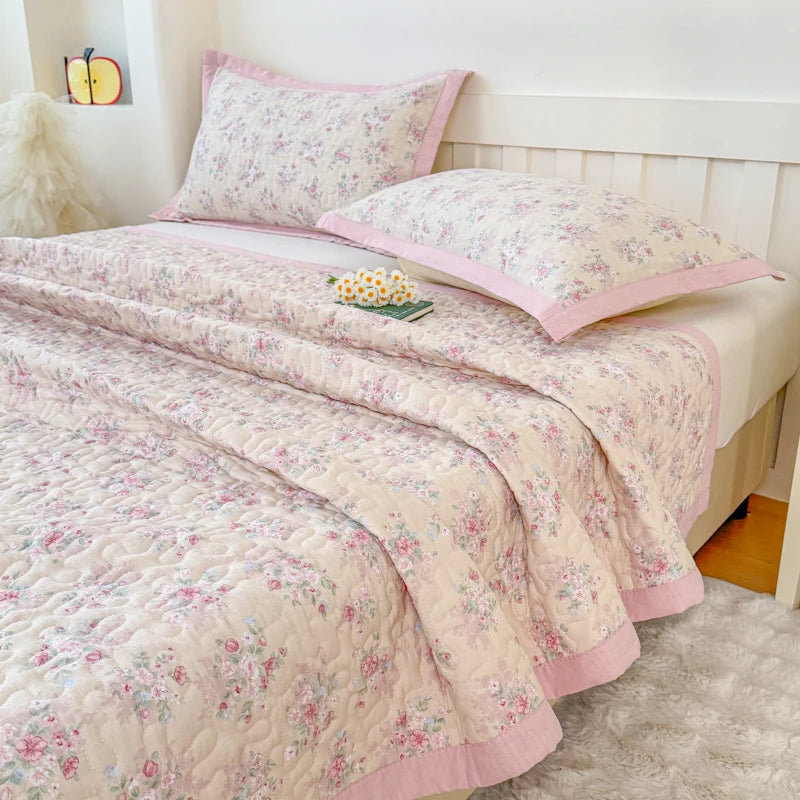 A pink floral quilted bedspread and pillows with a pink trim displayed on a white wooden bed frame.