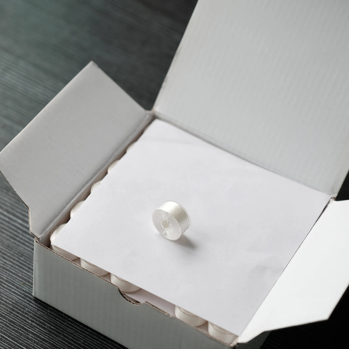 A white embroidery bobbin is placed inside a cardboard box for home embroidery machines.