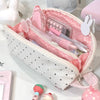 A pink polka dot shell-shaped pencil case with multiple compartments and a pink strap is shown on a white surface.