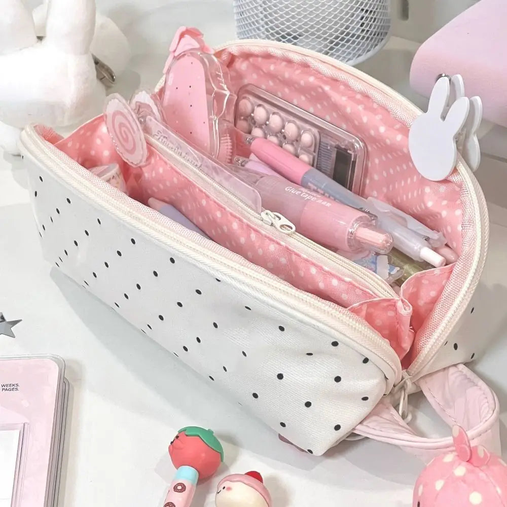 A pink polka dot shell-shaped pencil case with multiple compartments and a pink strap is shown on a white surface.