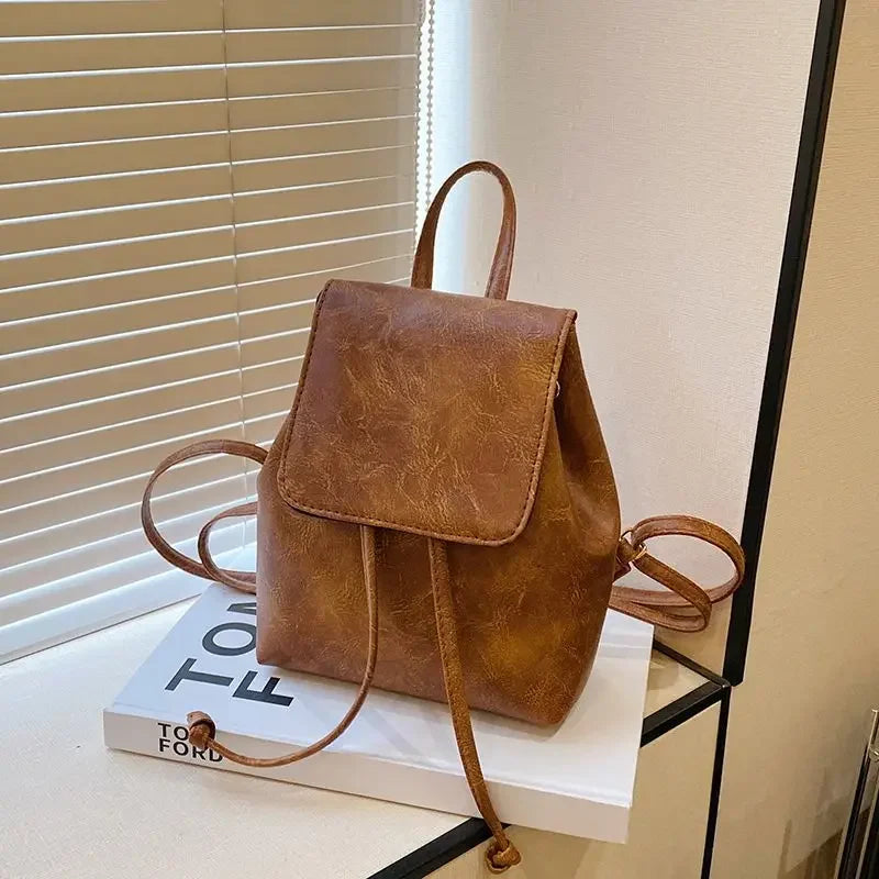 Petit sac à dos de mini en cuir marron avec cordon, exposé sur une surface blanche près d'un store.