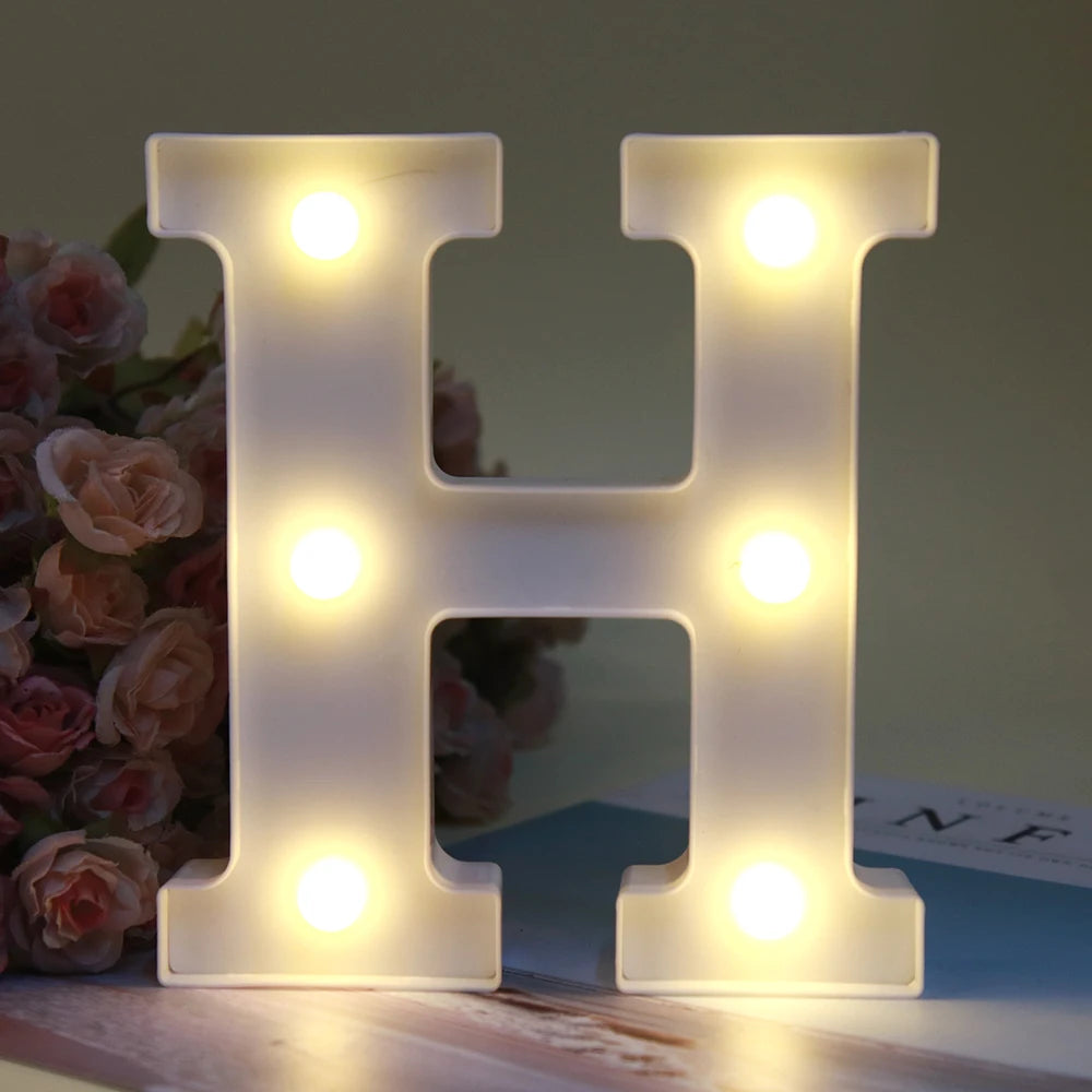 Lumière de nuit alphabet LED blanche en forme de H posée sur une surface en bois à côté de roses roses.