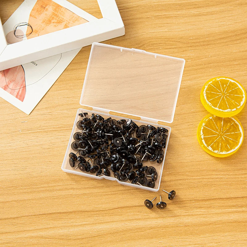 A clear plastic box containing a pack of black push pins and a glass of sliced lemon on a wooden surface.