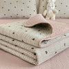 A stack of quilted fabric in pink and green gingham patterns is displayed on a bed, with a white stuffed animal in the backgr