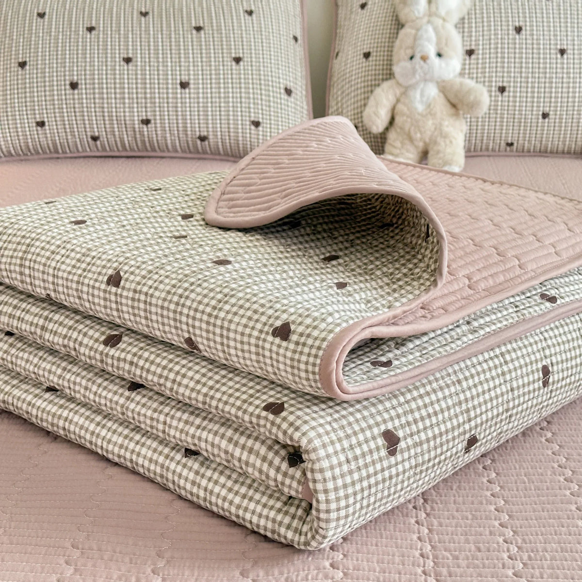 A stack of quilted fabric in pink and green gingham patterns is displayed on a bed, with a white stuffed animal in the backgr