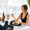 A woman is drinking red wine at a table set with black placemats and silverware.