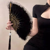 A hand holds a vintage folding fan with a black velvet surface and a lace-trimmed golden edge against a dark outfit.