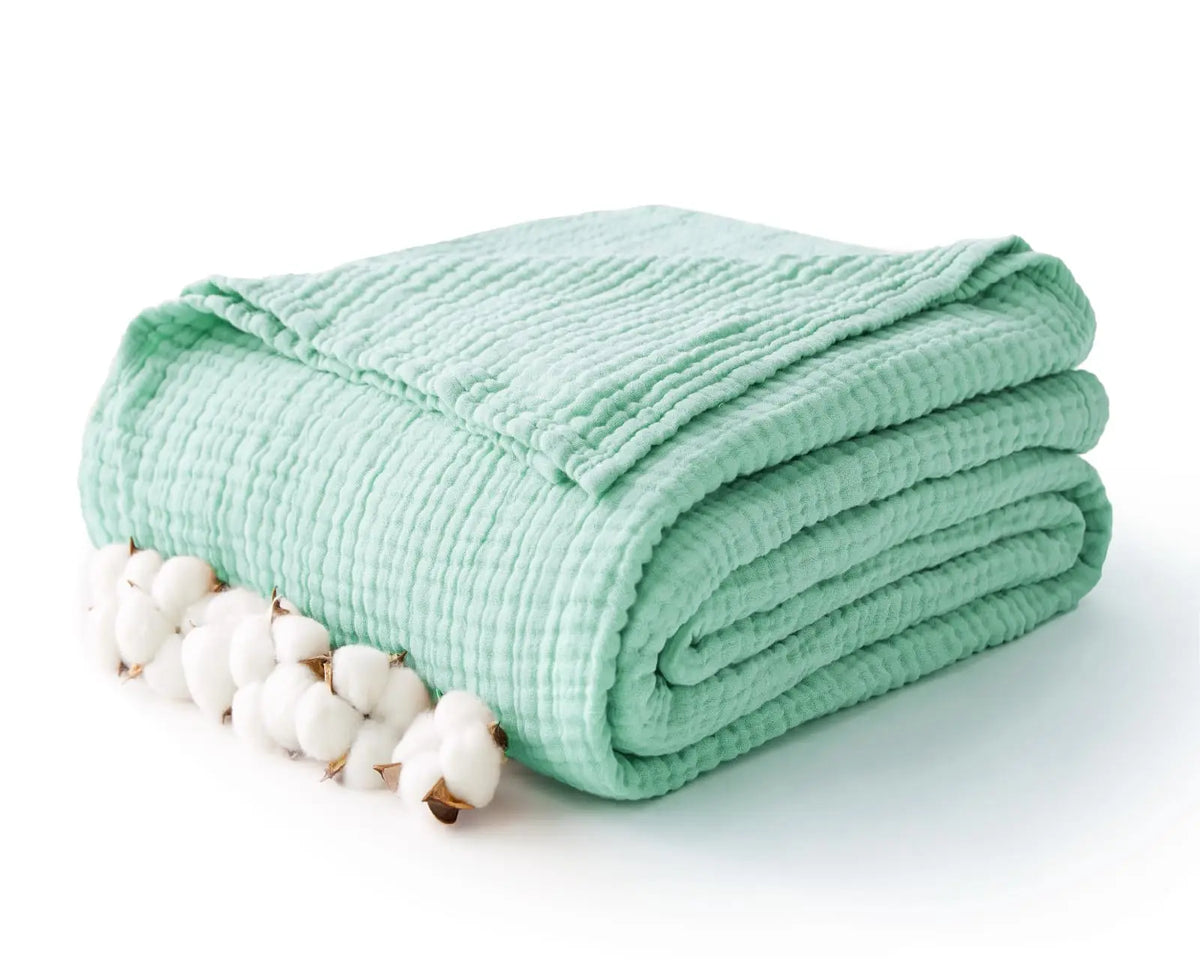 A light mint green soft muslin cotton blanket folded and accompanied by natural white cotton bolls.