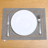 A white dinner plate and silverware are placed on a gray linen placemat with gold embroidery on a wooden table.