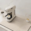 A white and gray coffee grinder sits on a beige counter mat in a bright kitchen setting.
