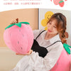 A woman is holding a pink strawberry plush throw pillow in a bright indoor setting.