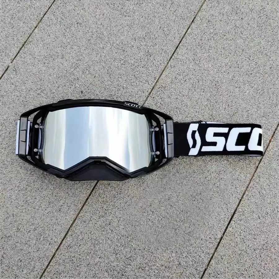 A pair of black SCOTT motocross goggles with a silver reflective lens and a black strap lying on a gray concrete surface.