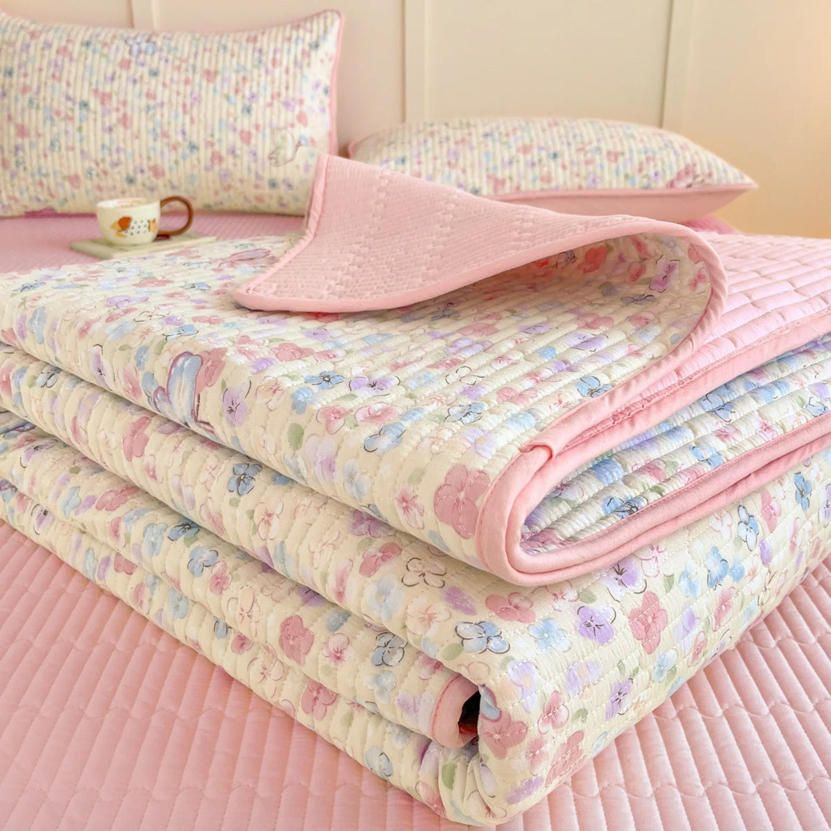 Pink floral quilted bedspread stacked with a pink dust-proof cover and a small cup.