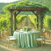 An elegant round satin tablecloth set up outdoors under a wooden arbor with greenery.
