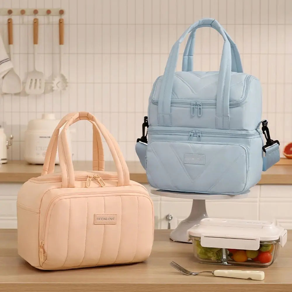 A light blue insulated lunch bag is displayed on a stand next to a pink reusable lunch bag.