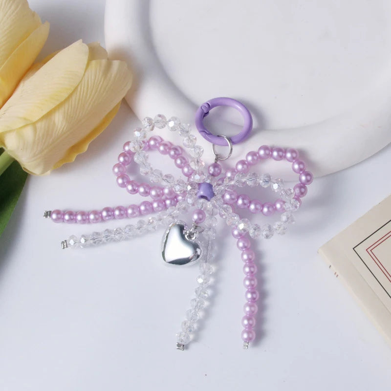A purple pearl bow keychain with a silver heart charm displayed on a white background next to a yellow rose.